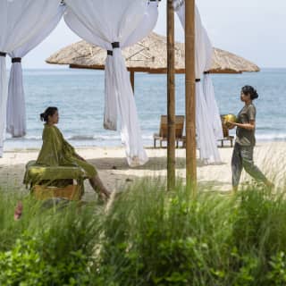 A staff member carries a fresh coconut to a woman in green, resting in a bamboo beach cabana with tied white curtains.