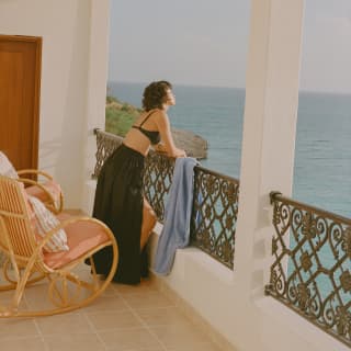On a balcony with wicker chairs, a woman in a black bikini top and skirt leans on the filigree railings, gazing at the sea.