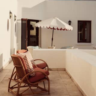 Two wicker chairs with plump orange cushions share a balcony connected to a terrace with a table, seating and white parasol.