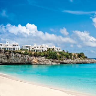 A two-masted sail boat enters Baie Longue's turquoise waters. At the top of a rocky outcrop rise the hotel's white walls