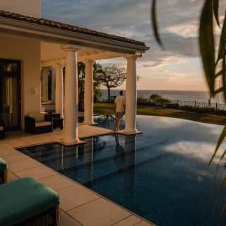 Seen from the far corner of the villa pool, a male guest leans against a terrace pillar in darkening light, watching sundown.