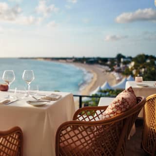 A dining table is set for two at the corner of the restaurant terrace with uninterrupted views of the sunset over Baie Longue