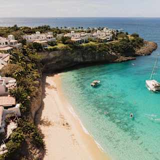 Four boats float in Baie Longue's sapphire waters, sheltered by a headland dotted with resort residences, in an aerial view.