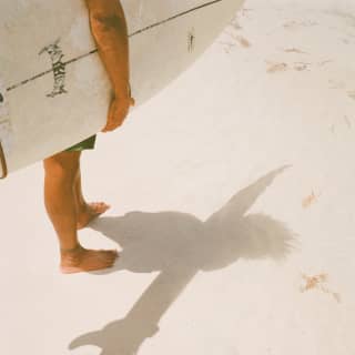 Close-up of a surfer's legs standing on the beach, casting a shadow his surfboard and wind-blown hair on the white sand.