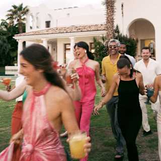 Six guests holding drinks and dressed up for an occasion laugh as they walk across gardens towards the camera, in close-up.