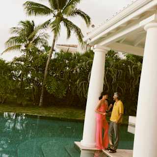 A man in a yellow shirt chats with a woman in a coral outfit by a pillar at the end of a terrace overlooking a still pool.