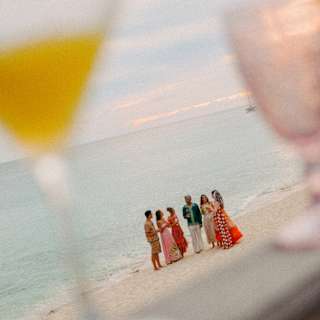 Six party-goers in bright clothing chat at the water's edge in an angled image, seen through cocktail glasses in soft-focus.