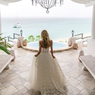 A bride in a flowing lace gown walks under the chandelier of the elegant hotel lobby as she looks out over the Baie Longue