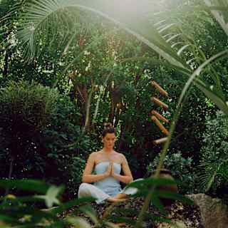 A woman in a blue strappy top and trousers meditates in the calm spa garden shaded from bright sun by lush foliage.