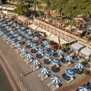 Parasols in blue and white candy stripes form neat rows along the rippled beach in front of the club bar, seen from above.