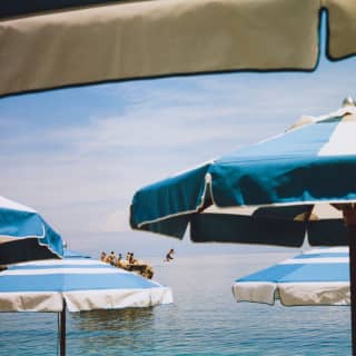 Action shot as a boy leaps into the sea from a rock where his friends sit in sunshine, seen through layers of blue parasols.