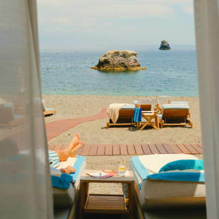View from a cabana over a sunbather and a pair of vacant beach chairs to the ocean, where two sea stacks jut from the blue.