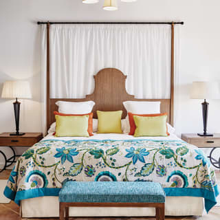 An archway frames a king bed with ornate bedhead, topped with white linens, bright cushions and a peacock blue floral throw