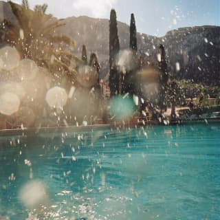 Sunlight glitters on tiny water droplets thrown into the air from a splash into a pool. Stone pines and palms reach for the sky