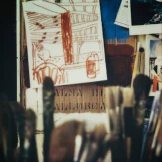 Sketches and images are pinned to an artist's easel. In the foreground, out-of-focus paintbrushes stand like tall grasses