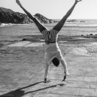 A man in white shirt and striped shorts performs a perfect cartwheel on stone paving. Behind a calm sea laps the shore