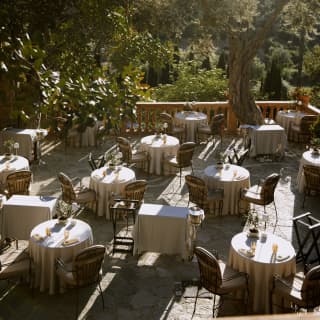 High angle view of the terrace where round white-linen tables are striped with light as it filters through the olive trees.