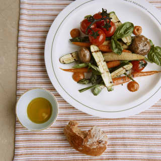 Looking down on a plate of courgette, corn, tomato and other grilled vegetables served with basil, dip and crusty bread.