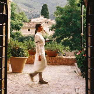 A white-shirted waiter in a long apron tied at her waist walks along the garden terrace, framed by an ancient stone arch