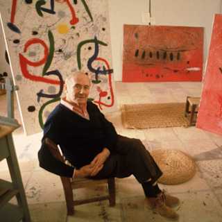 Photograph Joan Miró on a chair, crossing legs and hands, in a studio with a white abstract painting and two artworks in red.
