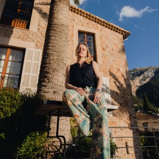 Ukrainian textile artist, Anastasiia Podervianska, sits on the edge of a table under a tall palm in the gardens of La Residencia