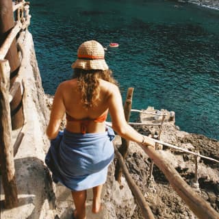 A woman wrapped in a blue towel sarong, holds onto a driftwood bannister as she makes her way down the Cala Deia steps