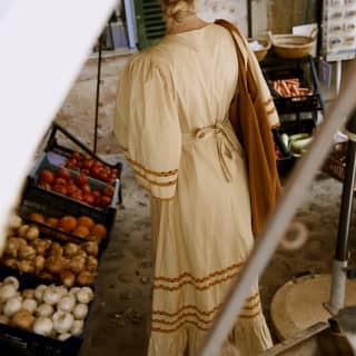 Seen from behind through a canopy, a blond woman in a white summer dress browses a vegetable stall in a corner of Deià.