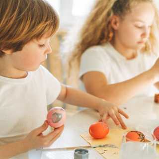 A young boy and girl, in white, with light hair, enjoy an Easter art activity at a table scattered with red and pink eggs.