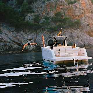 A man is captured mid-air diving from the Sa Brisa boat, moored in a bay, as a female friend watches from the deck.