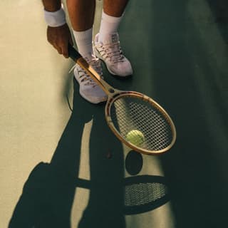 A man in tennis whites uses an old wooden racquet to bounce and scoop up a yellow ball. A low sun casts long shadows