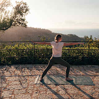 A woman in a white top and black leggings adopts the yoga warrior pose in gardens facing the sea, seen from behind.