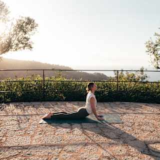 A woman in a white top and black leggings adopts the yoga cobra pose on a mat on a paved terrace with sea views.