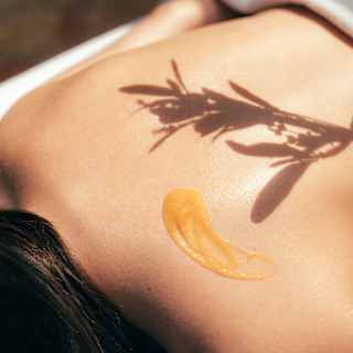 The shadow of an olive branch falls on the bare back of a spa guest dolloped with massage oil, in a close-up wellness detail.