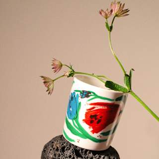 A white mug with colourful abstract designs is balanced on a black lava rock and a white stone, with a sprig of delicate pink flowers placed inside the mug, against a neutral background.