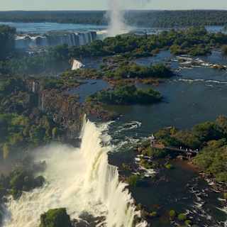 Aerial view of the Iguassu falls with creamy rapids cascading over the forested rocky edge, with more falls in the background.