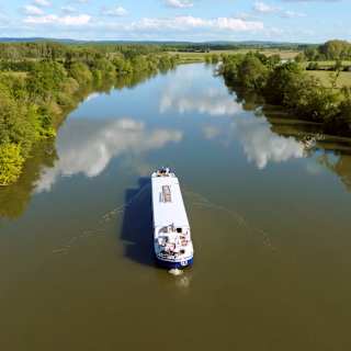 A blue and white barge cruises a waterway so calm it mirrors the clouds as sun illuminates verdant banks, seen from above.