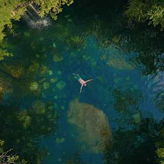 A person floats on their back in a clear, blue-green natural pool surrounded by lush, dense green trees, seen from above. Rocks and underwater features are visible beneath the surface.