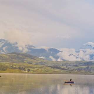 Snowy peaks rise on the horizon in a view over Laguna Huaypo, where a man tends to a boat on the still, green-tinted waters.