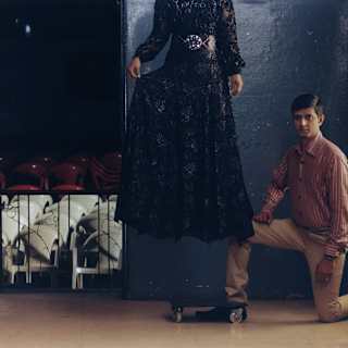A man in red-striped shirt and beige trousers kneels by a woman in a black lace dress, apparently floating above the floor.