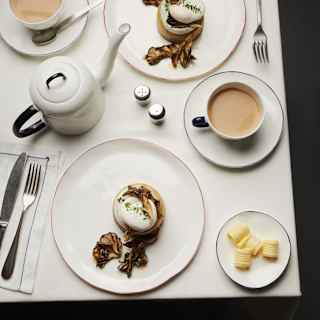 A white table set with two plates of eggs Benedict, two cups of tea with milk, a teapot, a small dish of butter, salt and pepper shakers, and cutlery arranged neatly.