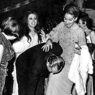 Film director Luchino Visconti stoops to help Maria Callas put her foot in a shoe in a historic black and white photograph.