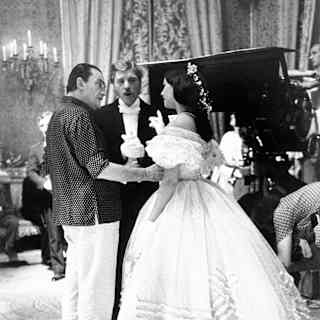 Visconti talks to Claudia Cardinale, who wears a white frilled dress, watched by Burt Lancaster on a film set, in monochrome.