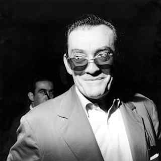 Film director, Luchino Visconti smiles as he is snapped in a suit with dark glasses, in a black and white close-up.