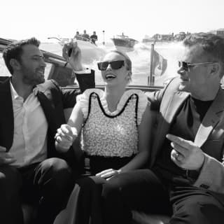 Black and white image of actors Ben Affleck, Jodie Comer and Matt Damon, dressed up and laughing together in a water taxi.
