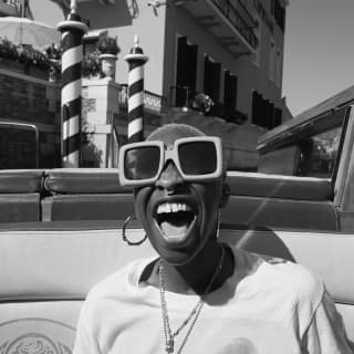Cynthia Erivo photographed in black and white, laughing in a water taxi, wearing large sunglasses, earrings and a pendant.