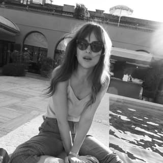 Dakota Johnson sits at on the pool edge with sunglasses, a drink, and one foot in the water, photographed in black and white.