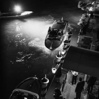In darkness, hotel staff wait as a late boat pulls up alongside the striped paline posts, guided by lamps, seen from above.