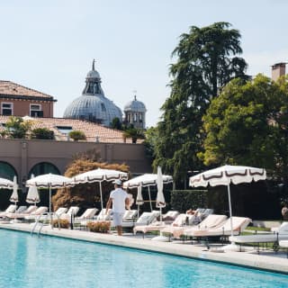 White sun loungers and parasols with brown-trim line the azure swimming pool, with the imposing dome of Le Zitelle behind.