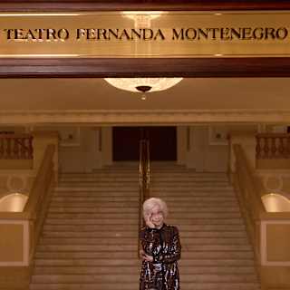 An elderly woman with white hair stands smiling in front of a grand staircase under a sign that reads Teatro Fernanda Montenegro. The elegant setting features ornate railings and warm lighting.