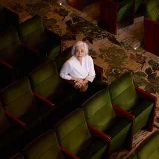 An elderly person with short white hair and a white shirt sits alone in a row of green theatre seats, looking up towards the camera. The surrounding seats are empty.